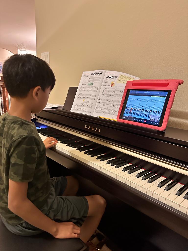 A boy plays a digital piano at home, with the score on the music stand and the Aquila piano diagram on a tablet beside it.